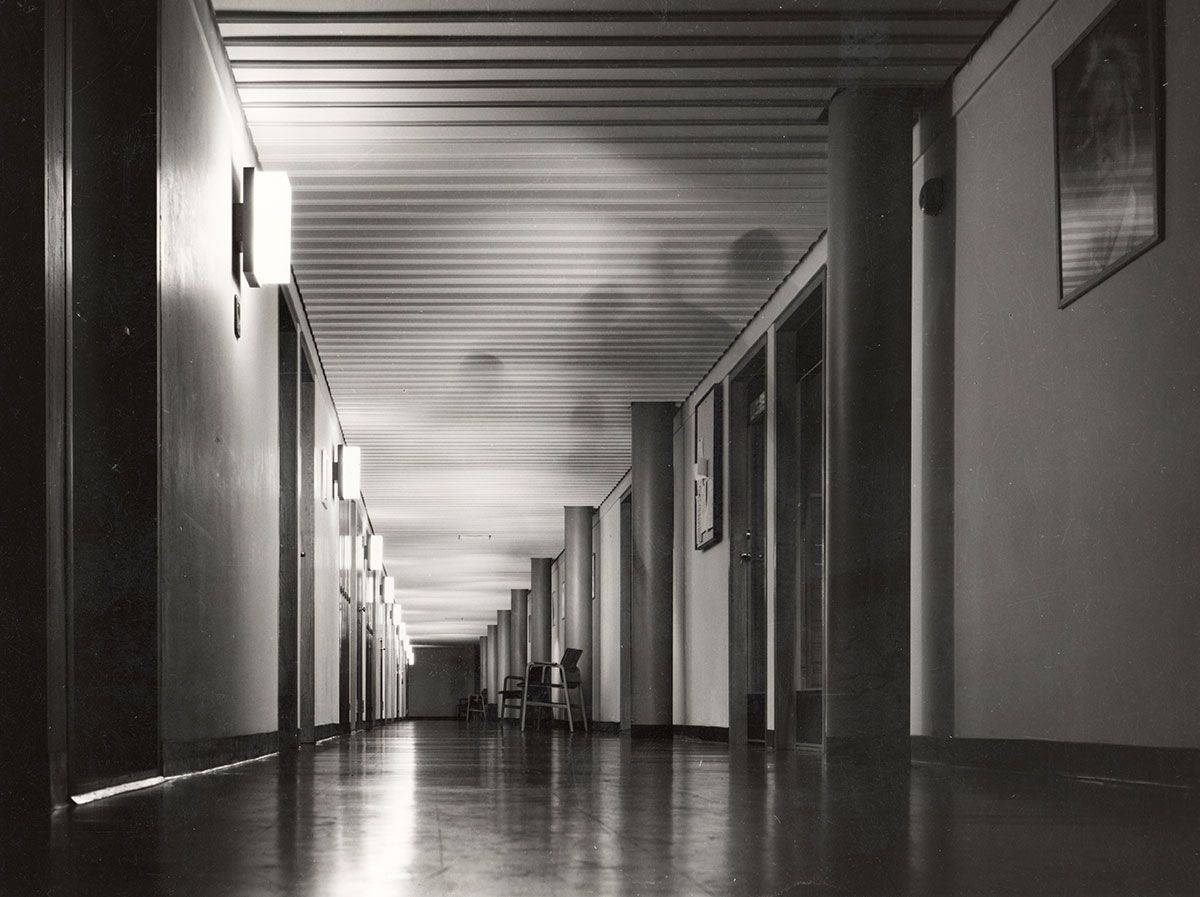 En av korridorene i Wergelands hus med linoleumsbelegg på gulvene og en prefabrikert elementhimling. De bærende søylene er rustrøde. På veggene henger lysarmaturer i plast tegnet av arkitektkontoret.&amp;#160;
1963-1965. Fotograf: Teigens fotoatelier.&amp;#160;